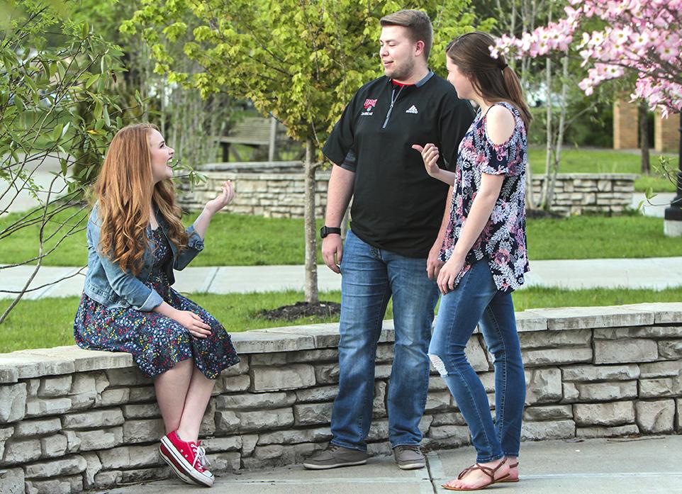 三个人, two standing and one sitting on a stone ledge, engaging in conversation outdoors near blooming trees and greenery.